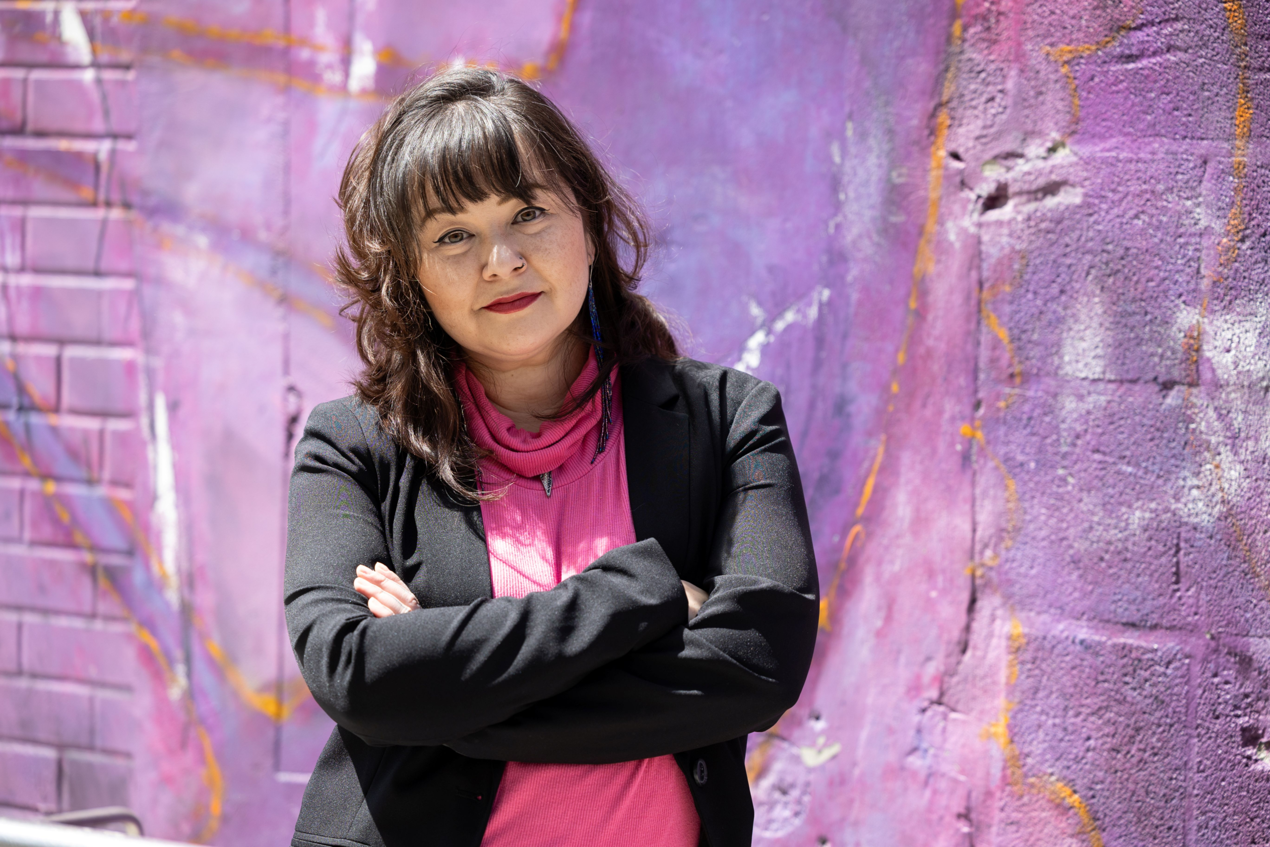 Victoria Davis stands in front of a fuschia & lavender Toronto street mural, arms crossed as she directly faces the viewer, as though to say: "Are you ready to change things for the better?"